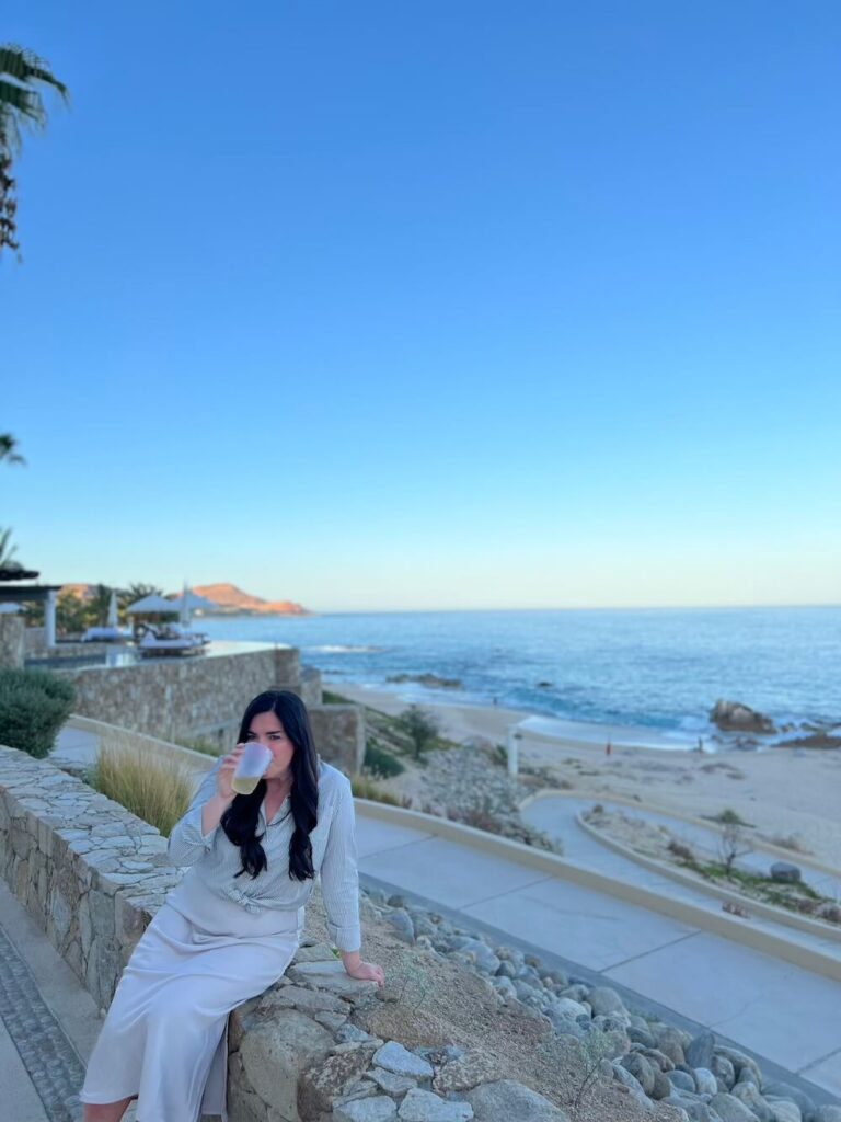 brunette woman drinking beverage from a cup with beach behind her