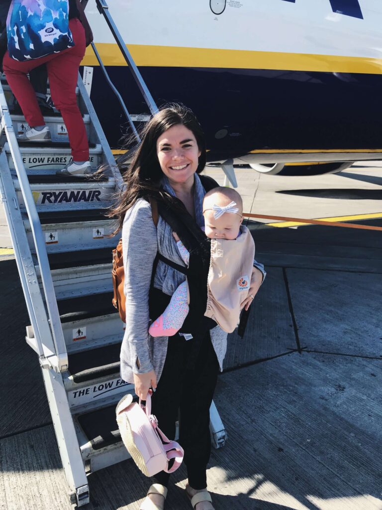 woman holding baby in a carrier standing in front of the steps to a plane