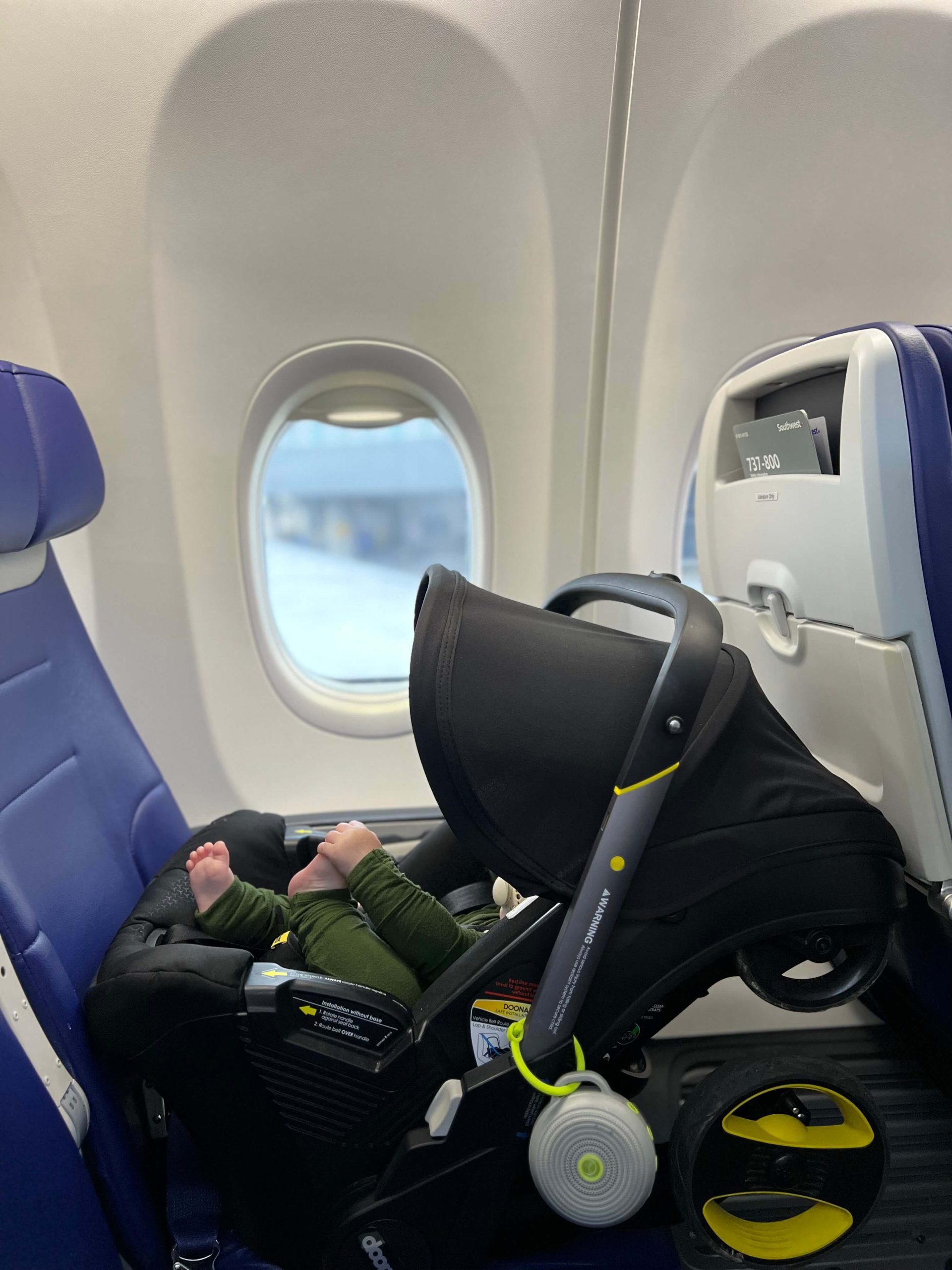 baby in a doona car seat with sound machine hanging off of the side in an airplane seat